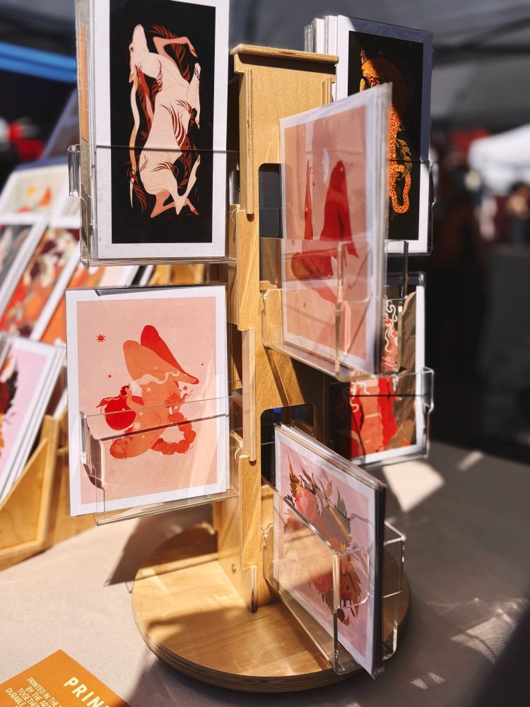 Photo of the Samantha Mash Illustration mini print rack. Morning sunlight hits the display with a selection of six mini prints in view on a tan table