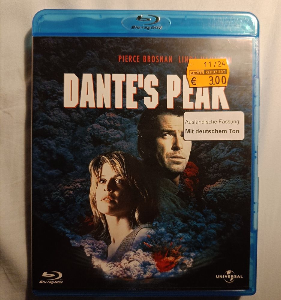 Das Cover der Blu-ray des Films Dante's Peak. Links unten sieht man Linda Hamilton, rechts etwas höher Pierce Brosnan. Brosnans Gesicht ist teilweise von einem Sticker mit der Aufschrift "Ausländische Fassung
 Mit deutschem Ton" verdeckt 