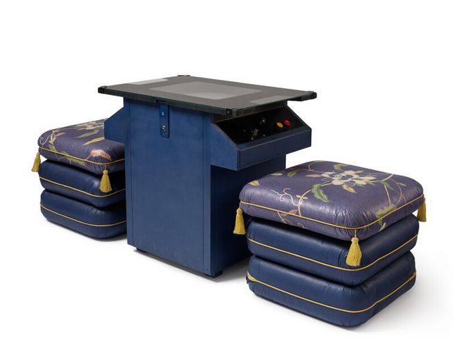 Screenshot der Sotheby's Homepage:
In der Mitte steht ein Pac-Man Tischspielautomat, ein kleiner Tisch dessen Tischplatte der Bildschirm des Spielautomaten ist. An einer Seite sind unter der Tischplatte kann man den Joystick und die Knöpfe des Automaten erkennen. Links und rechts sind so Kissenstühle, die Sotheby's wie folgt beschreibt: "associated ottoman style upholstered fabric stools"