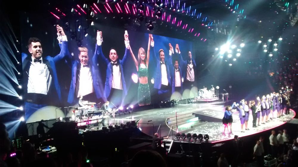 Ein Foto vom Taylor Swift Konzert in der Lanxess Arena 2019. Man sieht die Bühne, Taylor Swift steht in einer Reihe von Männern in blauen Smokings, sie bedanken sich für den Applaus. Auf einer Videoleinwand im Hintergrund sieht man die Szene nochmal größer