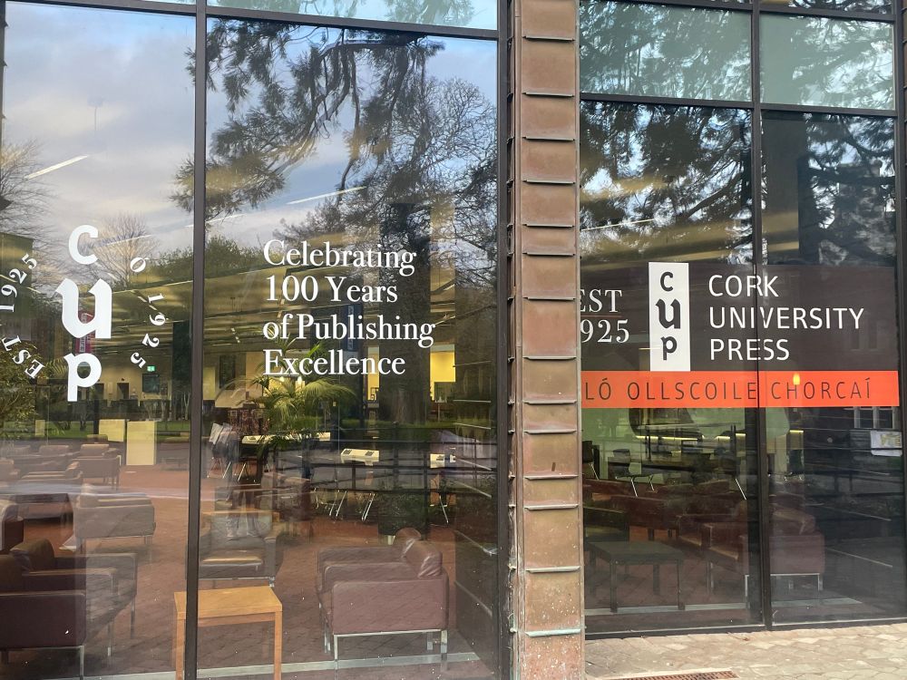 Photo of decals on a reflective window for the centenary of Cork University Press. Caption that says: Celebrating 100 years of Publishing Excellence. 
