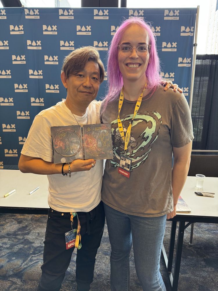 A picture of myself, with purple hair and a Final Fantasy 16 Zero t-shirt posing with Yoshi-P while he holds a signed steelcase of Final Fantasy 16.