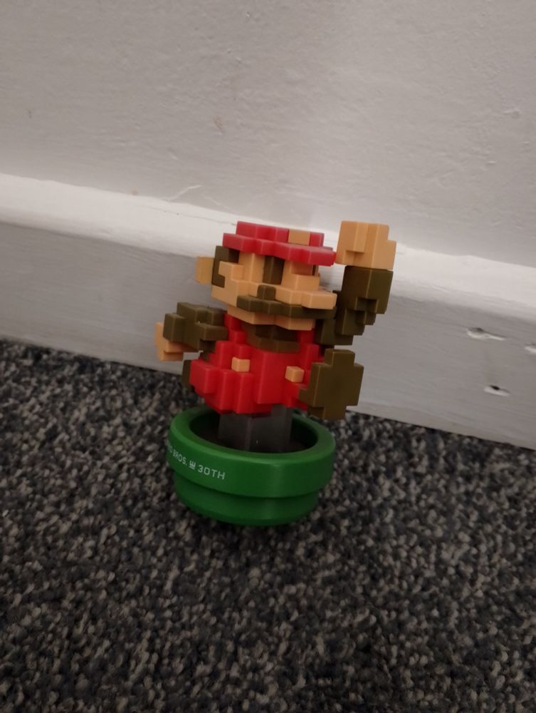 A picture of the 30th anniversary 8-bit Mario (classic colours) amiibo slanted against a white wall.