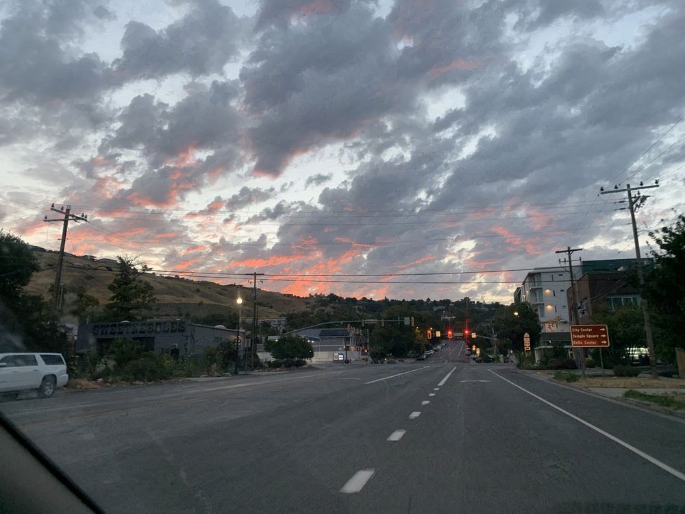 A photo of the sky just before sunrise, with orange light illuminating the bottom of gray clouds, above a forested hill. In the foreground, a street and streetlights.