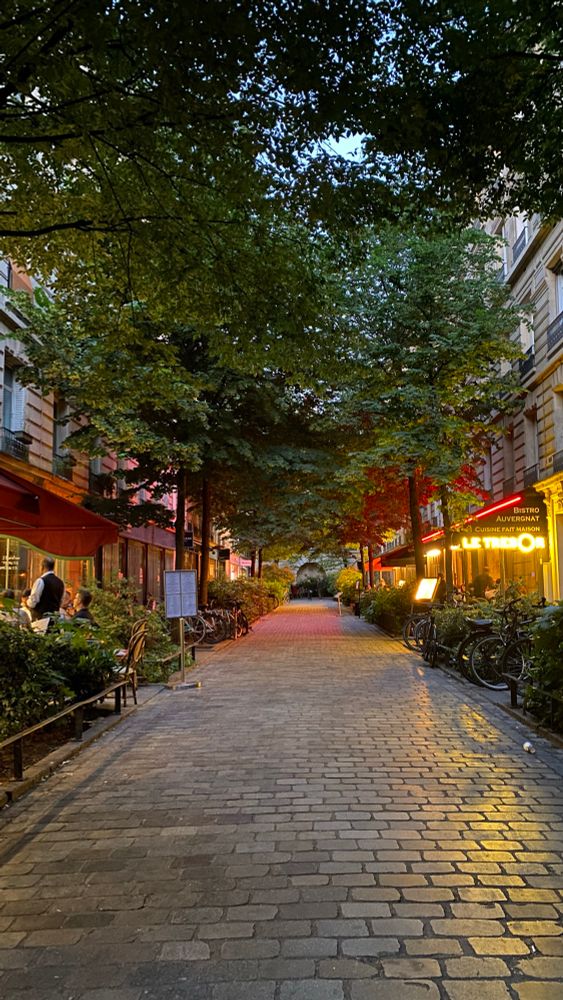 Softly-lit paved pedestrian street lined with trees and outdoor seating for restaurants at dusk in Le Marais, Paris