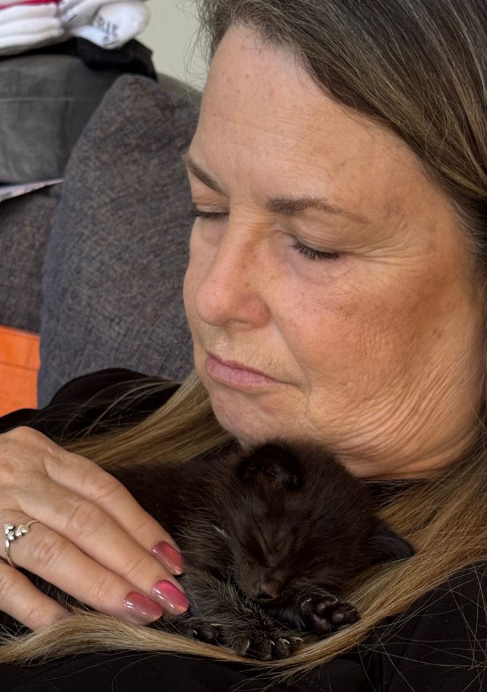 A photo of my wife, wearing a black sweater, holding a tiny black kitten. Both have their eyes closed and appear to be asleep. 