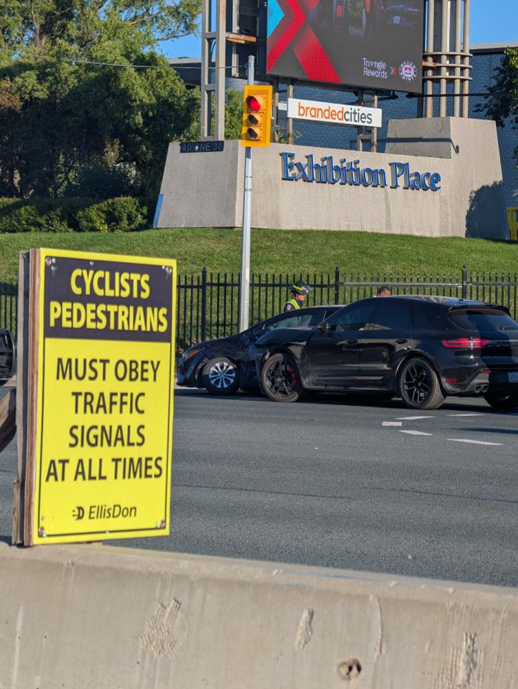 Photo of a sign on the multi-use path on Lakeshore Blvd in Toronto which says, "Cyclists Pedestrians must obey traffic signals at all times". In the background, two cars have collided in the intersection with significant damage to both.