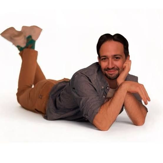 A photo of lin manuel miranda, laying on his stomach and smiling with his feet kicked up in the air like a teenage girl in a 90s sitcom writing in her diary or perhaps waiting on a phone call from her crush