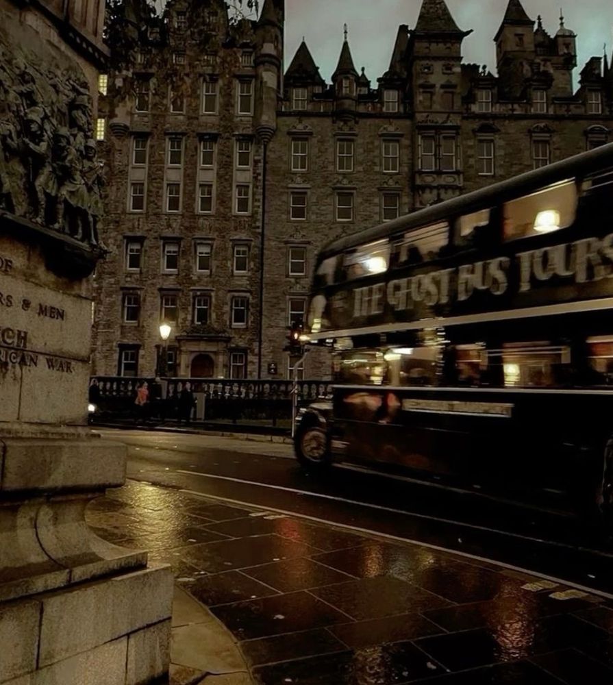 A bus speeds by old stately stone buildings, it's dusk out so things are only illuminated by the glow of streetlights, and the street and pavement are wet so it must have just stopped raining. 