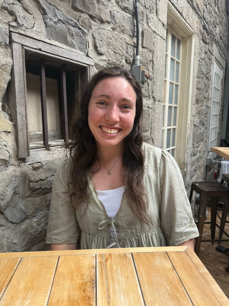 a brunette woman (me) sitting at a wooden table with a cobblestone wall in the background. i am wearing a sage green shirt with a white tank top underneath 
