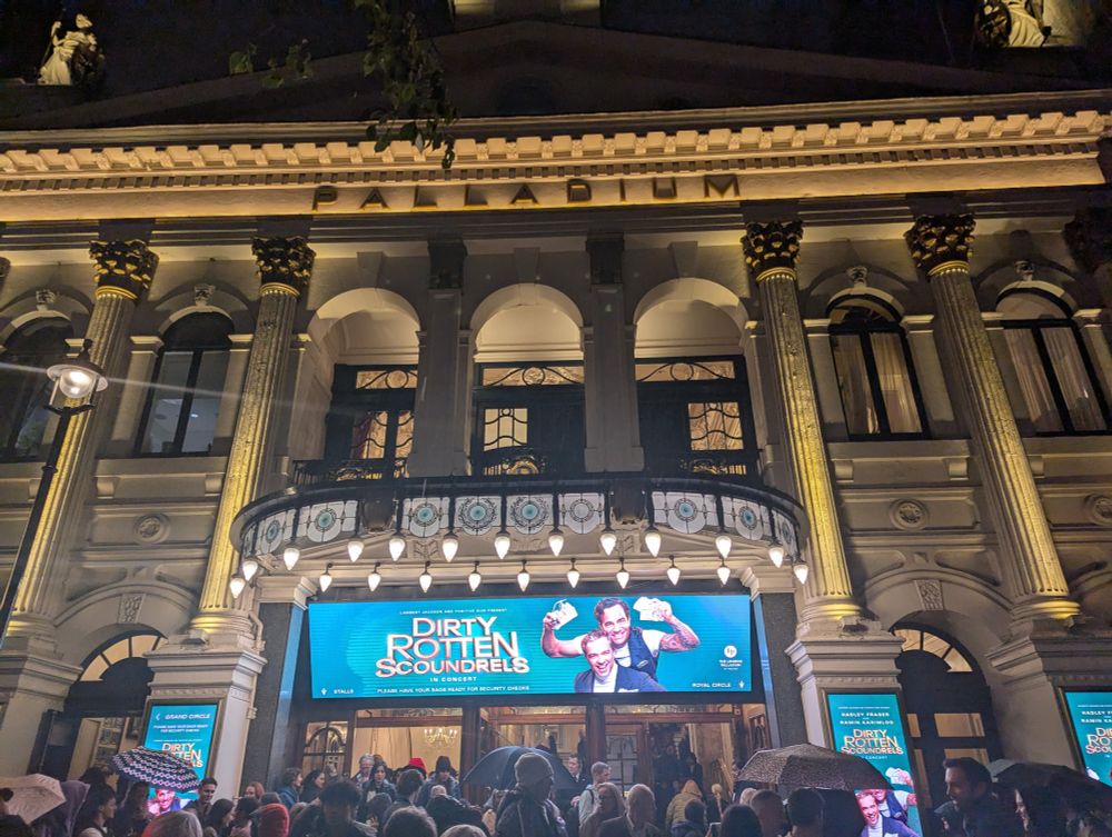 An image of the London Palladium theatre at night showing a large poster promoting Dirty Rotten Scoundrels musical showing the actors Hadley Freeman and Ramin Karimloo. There is a crowd outside the theatre with umbrellas as it's raining. The theatre is floodlit.