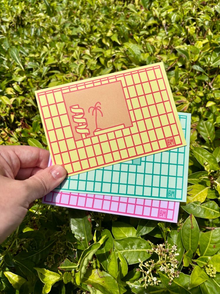 Three postcards held against a bush. Each card has an illustration of The Atlantis building in one of three colors: red/yellow, green, or pink. 