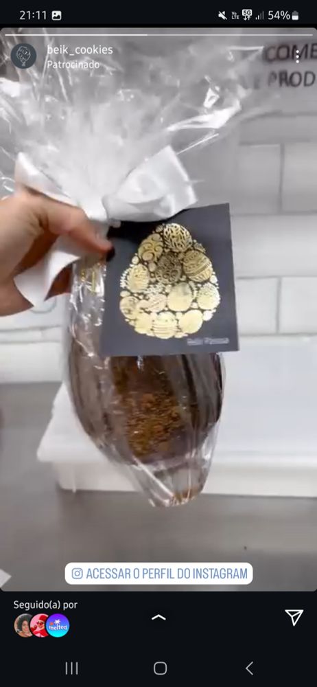 Print de um stories patrocinadk do instagram, da pra ver uma mao de uma oessoa branca segurando um ovo de pascoa com embalagem transparente, dentro um ovo de chocolate sem muita nitidez devido ao plastico. Um laço de fita branca amarra a embalagem, com um cartao preto com decoracao dourada escrito (em baixa nitidez) Feliz Páscoa