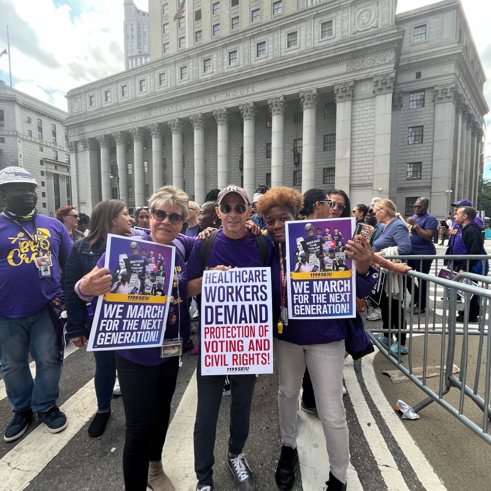 A group photo of three 1199ers at the March On Wall Street on August 28, 2025. They are wearing purple and gold union shirts. They are holding signs that read, 