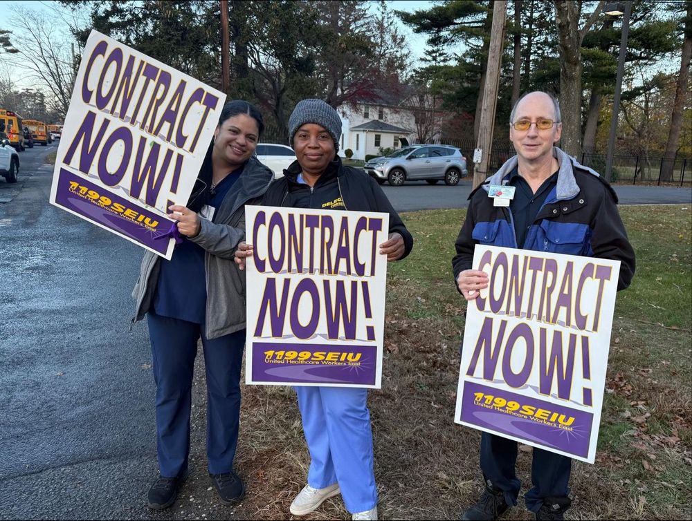 1199ers from Maria Regina Nursing Home in Brentwood, Long Island, New York picketing for a fair union contract. They are holding signs that say "Contract Now!" with other purple and gold union swag. 