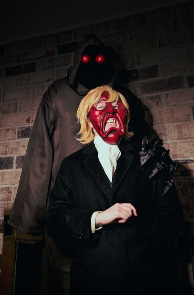 Two sinister figures in front of a brick wall. One, taller, is cloaked with glowing red eyes. Their taloned hand is on the shoulder of the other, who wears a dark suit, white cravat, and a grotesquely frowning red mask.