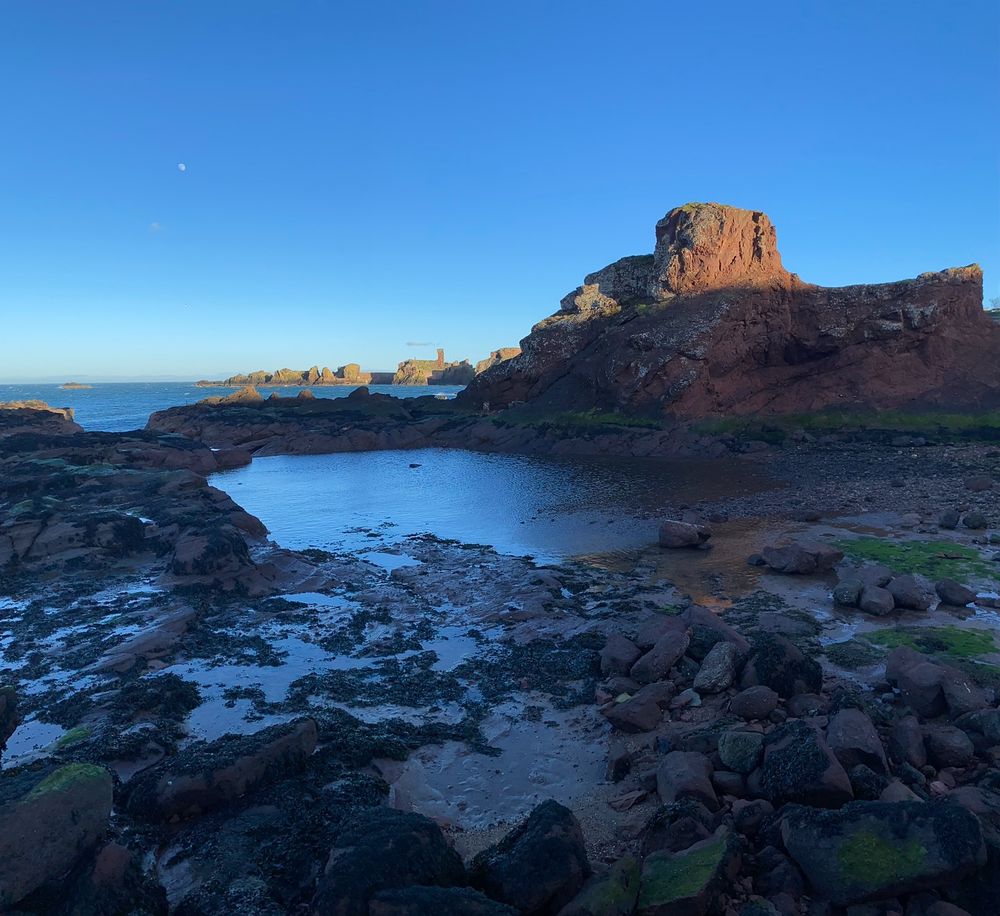 Belhaven Bay, Scotland. A moody blue rockpool reflecting a moody blue sky, looking out to sea past burnt orange rock formations. A tiny partial moon hangs in the sky.