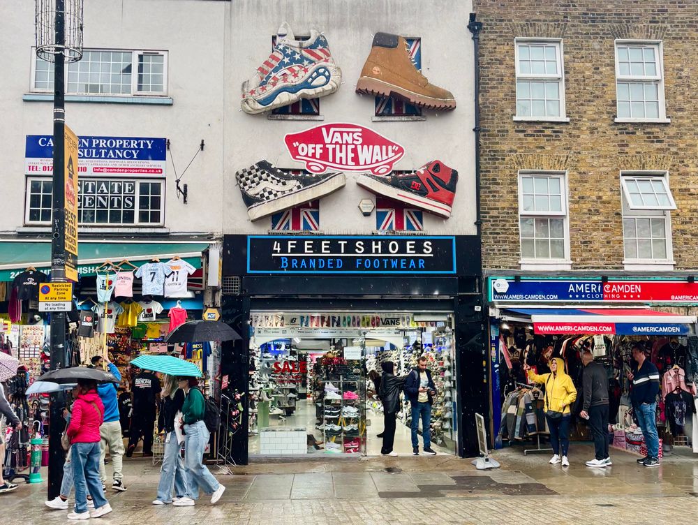 People with umbrellas walking past shops. One shop has giant shoes up on the wall above.theres some tshirts hanging up on a shop to the left. People sheltering from the rain in the doorway of shop on the right.