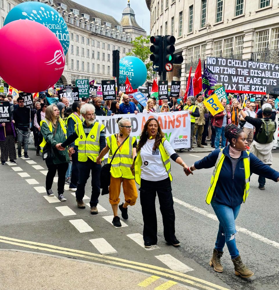 People in high vis clothes holding hands leading lots of people on protest march, lots of banners and large balls with writing on.buildings in the background.