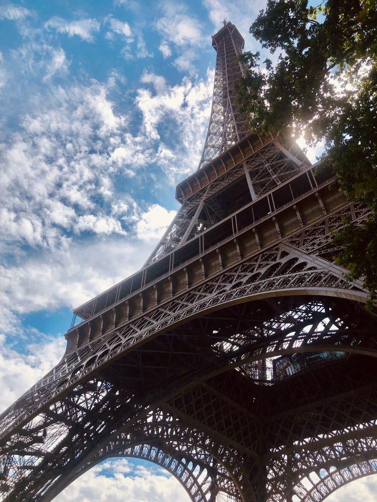 Looking up at tall steel tower, straight lines and arches. clouds in the sky and a bit of tree.