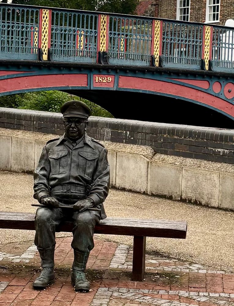 Statue of a man in army uniform sitting on a bench. Behind him is a bridge with the date 1829 on.