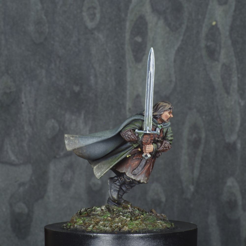 Photo d'une figurine peinte de Aragorn du seigneur des anneaux. Il est debout en train de courir son épée tenue à deux mains sur son côté droit. Il est vêtu d'un surcot en cuir brun sombre, d'un manteau vert et d'une cape verte desaturée. La scène se passe en forêt, le sol est brun et vert. 