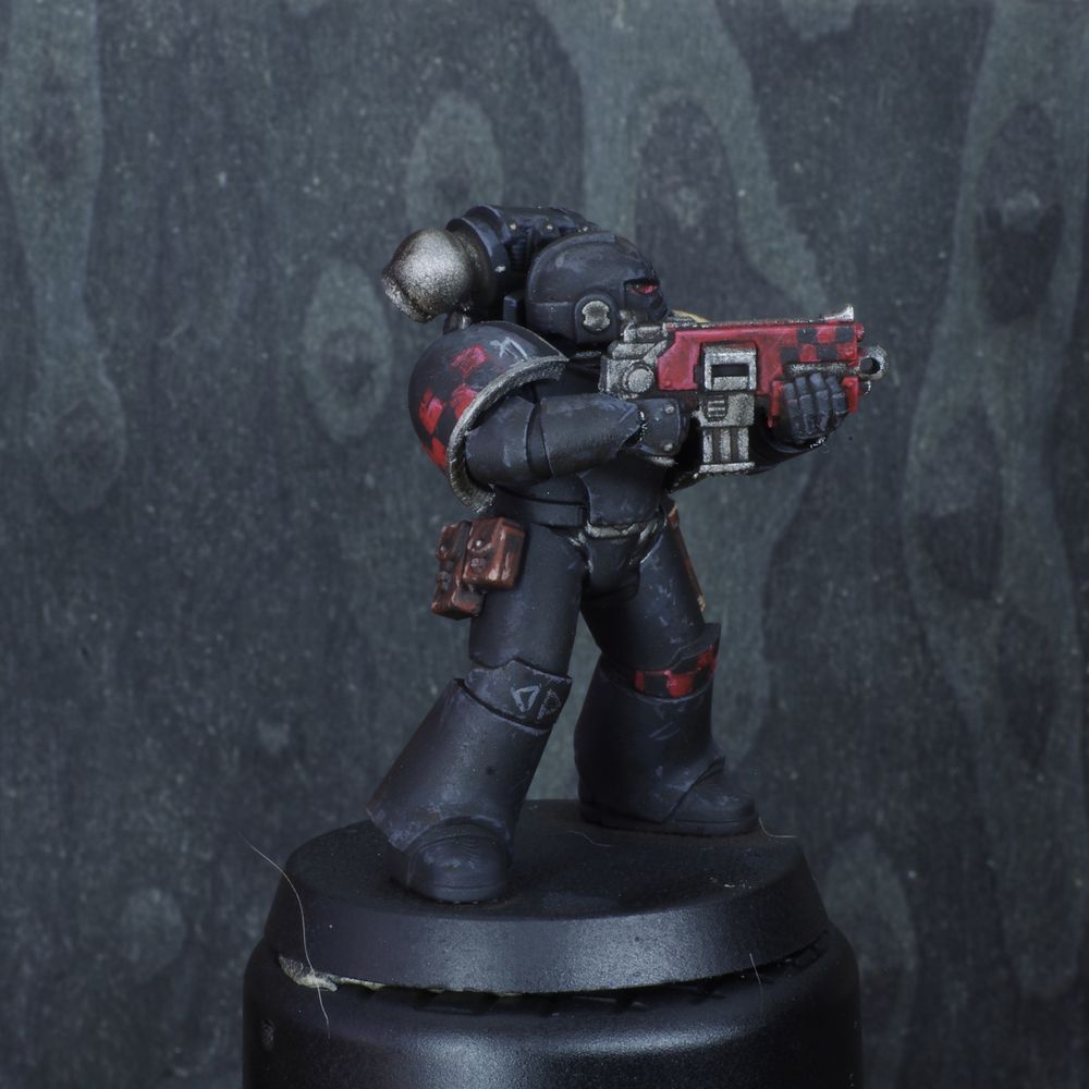 Photo d'une figurine de space marine de warhammer 30000 l'hérésie d'horus. Iel est un Dark Angel, son armure est noire et usée, son épaule gauche ets couleur os. Il y a des damiers rouges et noirs sur son genou gauche, sur son épaule droite et sur le bolter.