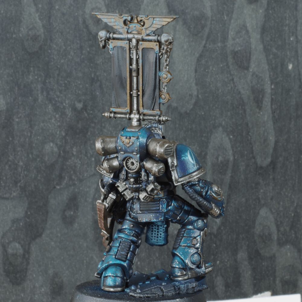 Photo de la figurine d'un Centurion Space Marine, iel est debout sa main droite posée sur son genou et sa main gauche tient un Bolter combiné. Son armure est richement décorée, c'est un modèle Mk2, son casque est un modèle Mk6 custom Alpha Légion. L'armure est de couleur bleutée avec des ombres violettes, les bordures sont argentés. Une grande bannière est fixée sur son dos.
