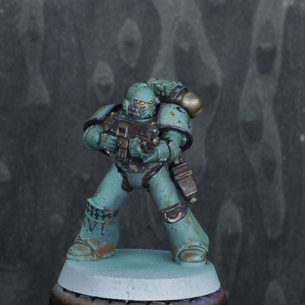 Photo d'une figurine de space marine de warhammer 30000 l'hérésie d'horus. Iel est un Sons of Horus, son armure est verte désaturéee, elle est sale et usée. Il y a des détails noirs sur l'armure. 