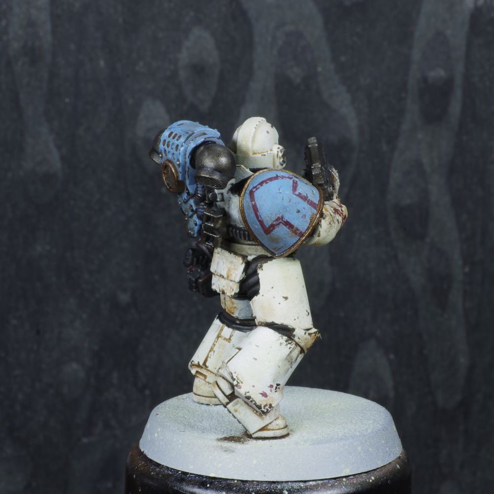 Photo d'une figurine de space marine de warhammer 30000 l'hérésie d'horus. Iel est un World Eaters, son armure est blanche avec des motifs rouges, elle est lourdement usée et sale. Ses épaules sont bleues désaturées.