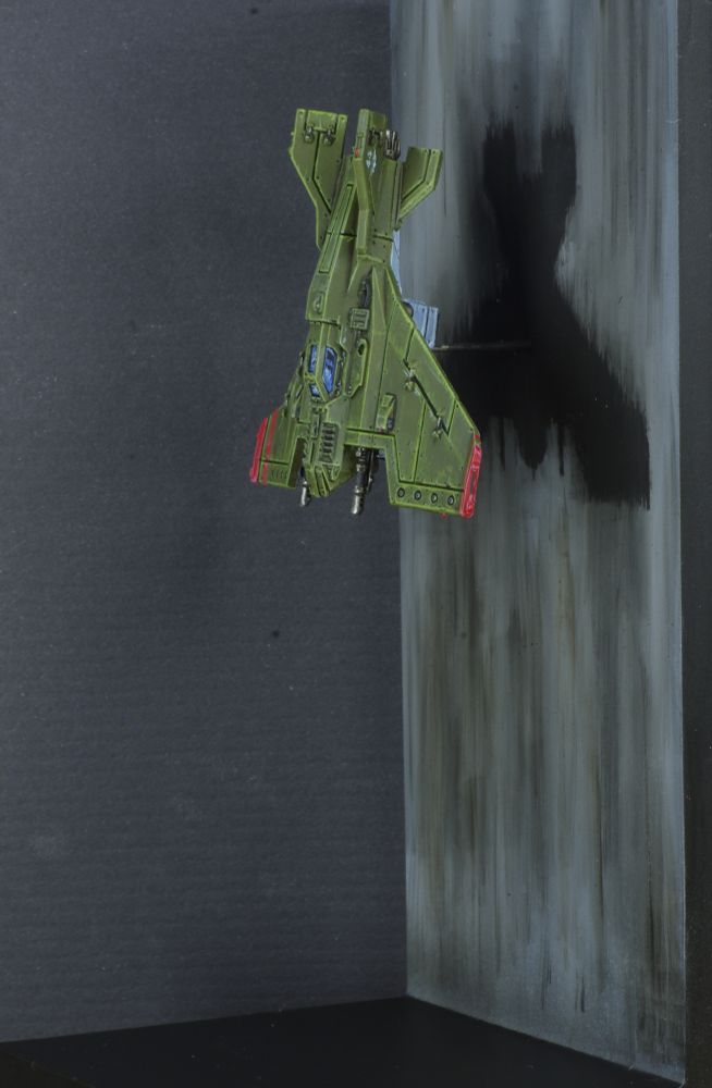 Photo d'une figurine d'un avion en chute libre devant un mur. L'avion est vu de côté, c'est est un chasseur Lightning Voss de warhammer 40 000, ses ailes sont une aile delta inversée. L'avion est peint en vert, au bouts des ailes sont appliqués deux bandes rouges. 