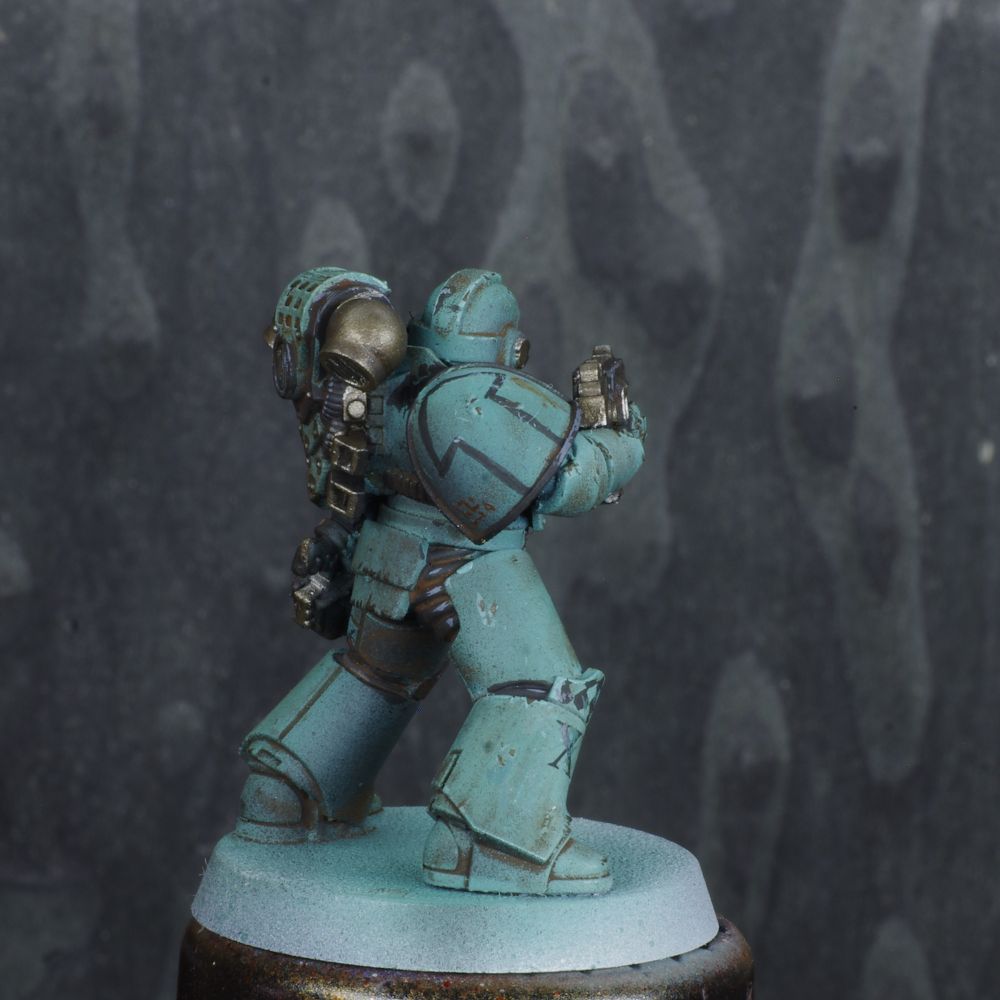 Photo d'une figurine de space marine de warhammer 30000 l'hérésie d'horus. Iel est un Sons of Horus, son armure est verte désaturéee, elle est sale et usée. Il y a des détails noirs sur l'armure. 