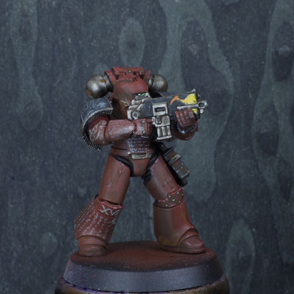 Photo d'une figurine de space marine de warhammer 30000 l'hérésie d'horus. Iel est un Word Bearers, son armure est rouge brune, elle est couverte de tunes blanches et de motifs ésotériques. Une flamme est dessinée sur son bolter. 