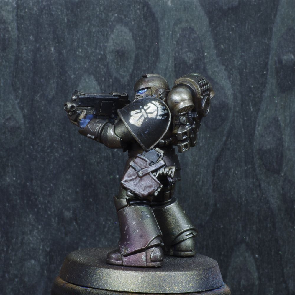 Photo d'une figurine de space marine de warhammer 30000 l'hérésie d'horus. Iel est un Iron Hands, son armure est un métallique sombre avec des reflets verts et violets. Il y a des motifs blancs dessinés un peu partout sur son armure. 