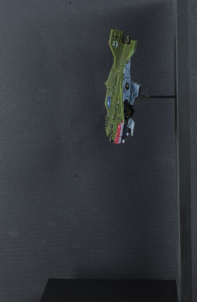 Photo d'une figurine d'un avion en chute libre devant un mur. L'avion est vu de côté, c'est est un chasseur Lightning Voss de warhammer 40 000, ses ailes sont une aile delta inversée. L'avion est peint en vert, au bouts des ailes sont appliqués deux bandes rouges. 
