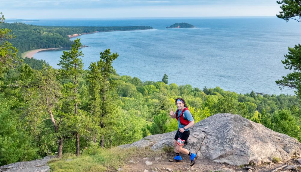 A picture of me running on top of a peak with forrest and Lake Superior below me. There is a sandy beach off the lake below. 
I'm wearing running gear and a running vest, with poles in a quiver on the back, and a running number on my shorts. I'm giving the Shaka symbol with my hand. 