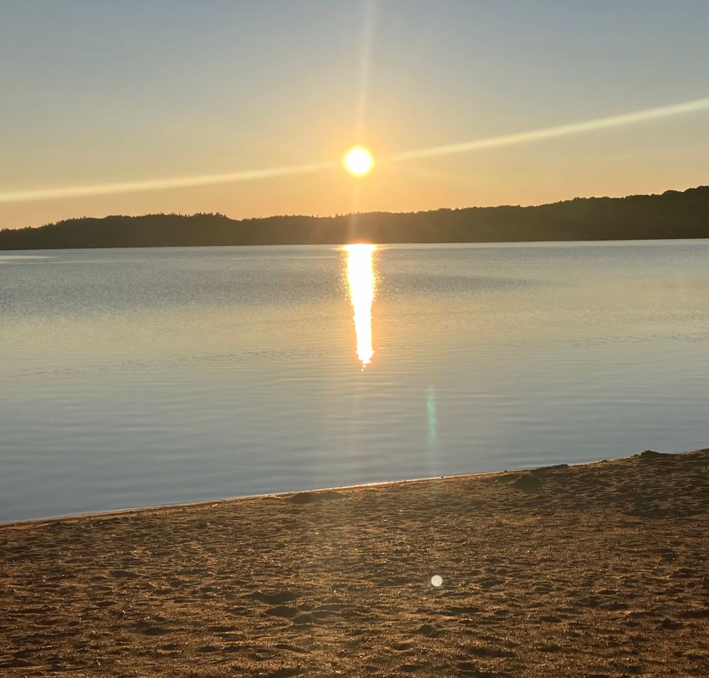 Sun setting over a lake with a sandy beach and hills in the background 