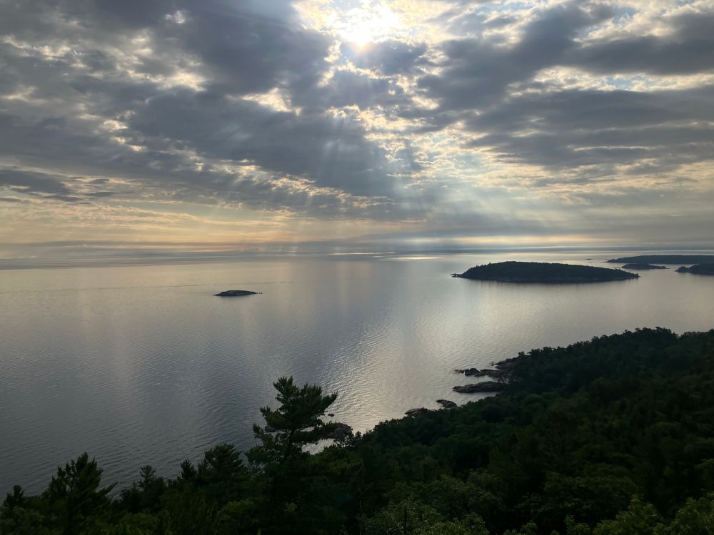 Picture of Lake Superior from the hills on the shore. Foreground has some trees and the sky is cloudy with the sun’s rays poking through. A couple little islands are offshore