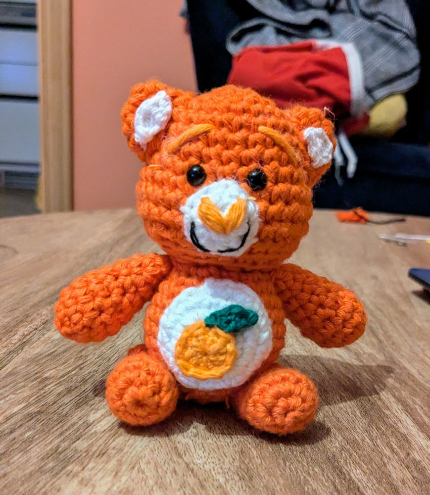 a care bear style bear, crocheted by OP, inspired by jim from our flag means death. they are dark orange with a white muzzle, ear inners and tummy patch. there is an orange with a small green leaf on the tummy patch. 