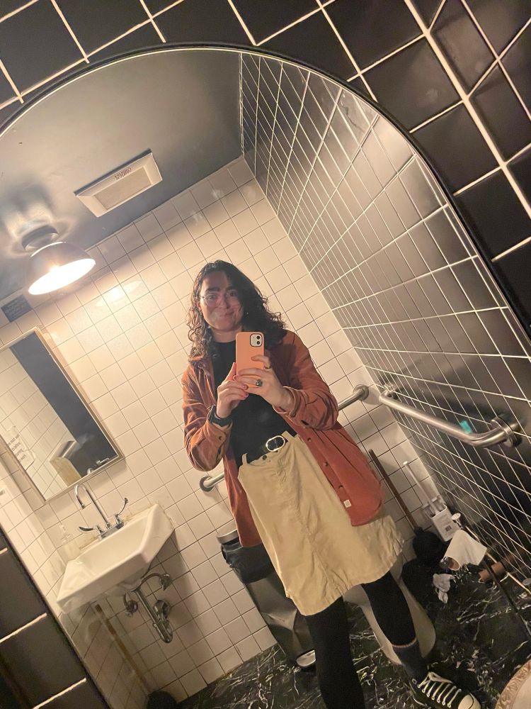account holder in a dirty mirror in a bathroom, wearing tights, converse, a tan knee-length corduroy skirt , a black mock neck shirt and brown corduroy jacket. she smiles 