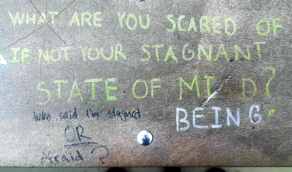 A park bench where someone has written "What are you scared of if not your stagnant state of mind?" Someone else has crossed out "Mind" and replaced it with "Being". Another person has written "Who said I'm stagnant or afraid?" beneath.