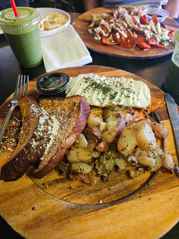 Appetizing photo of a diverse breakfast plate including seasoned potatoes, an over easy egg over chilaquiles and carne asada and French toast. In the background is a green smoothie as well as a second plate of chilaquiles in red sauce.