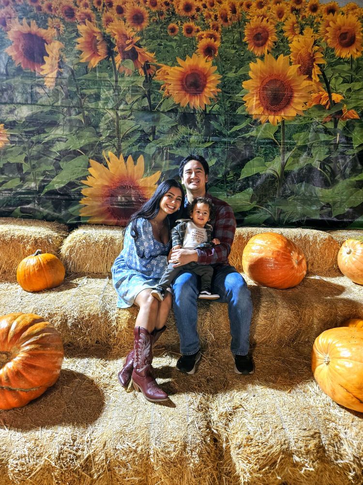 Family of 3 sitting on a hay bales surrounded by pumpkins. The backdrop is of a sunflower field. 