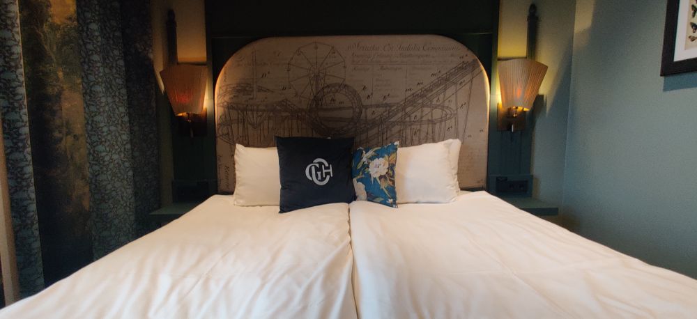 A photo of a super king size bed with white linens and pillows with floral print and with the hotel's monogram. The headboard has a diagram of an olf fashioned roller coaster, with fan shaped lampshades on either side.