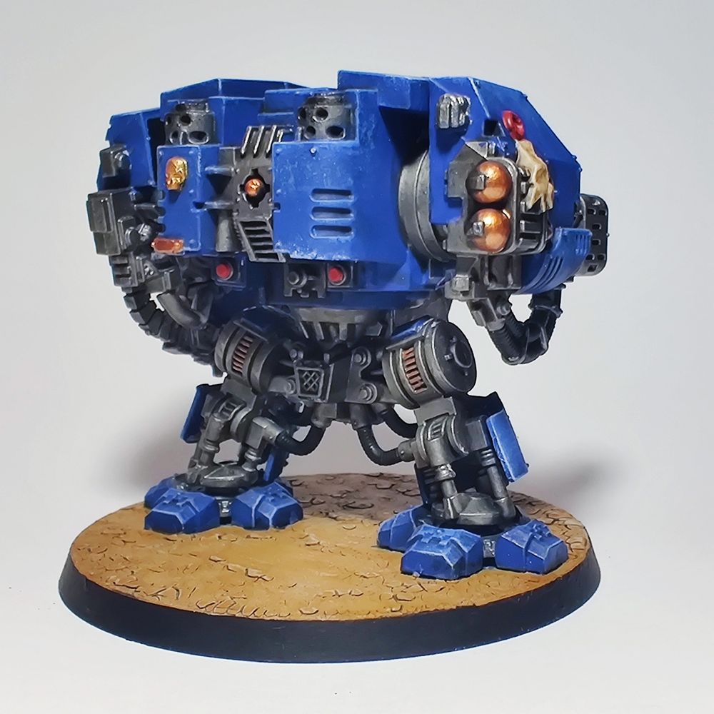 A Ultramarine Dreadnought miniature from the Assault on Black Reach set. It is a rear view standing at a three-quarter angle facing right. It is a bulky blue mecha with a gun arm and a claw arm. It is standing on crackled earth.