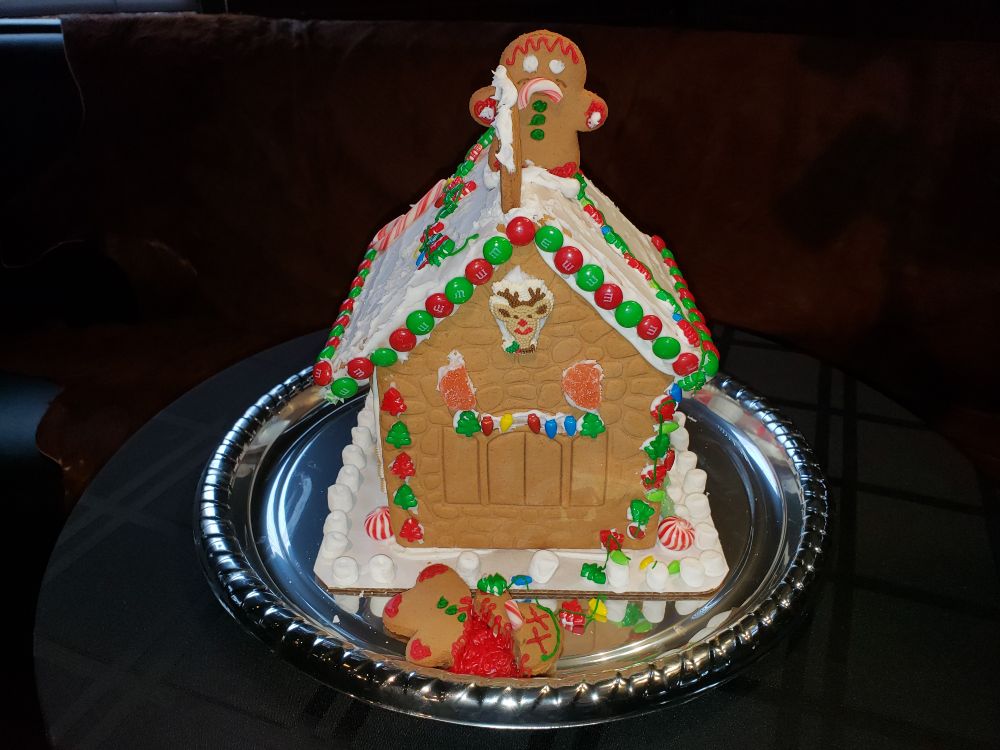 Photo of a ginger bread house with a white frosting roof, trimmed with red & green m&ms, Christmas trees, and holiday lights. On top is a gingerbread man with a candy cane frown, looking down on a gingerbread man lying on the ground, his head separated from his body, red frosting Xs for eyes, and red frosting in the gap between his body and head (as a substitute for blood).