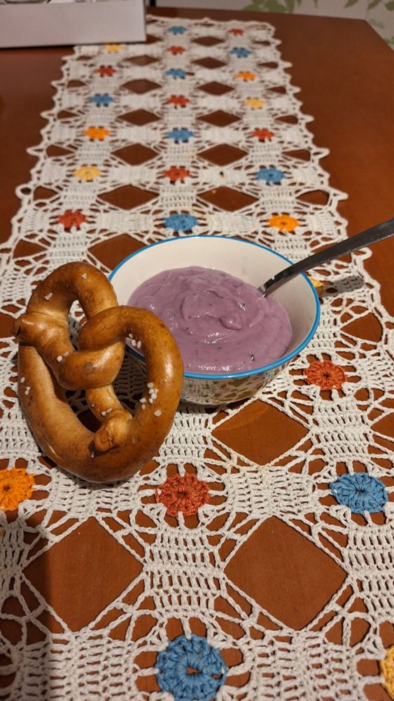 Fjólublá blómkálsúpa í lítilli skál og mjúk brauðkringla á eldhúsborði. Á borðinu er heklaður hvítur og blómóttur dúkur / Purple cauliflower soup in a small bowl and a soft prezel on a table. The table has a crochet white and flowery table runner.