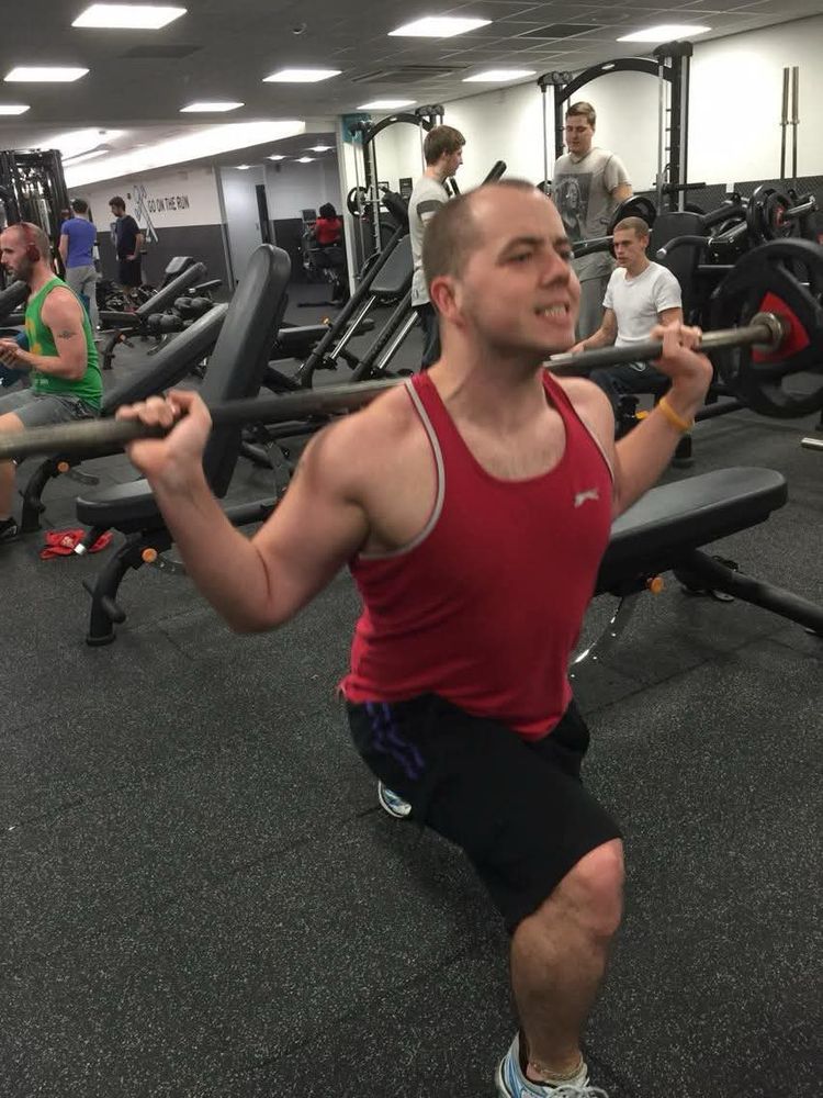 A gym venue.
A guy with a red vest top on, doing forward lunges with a barbell on his shoulders 