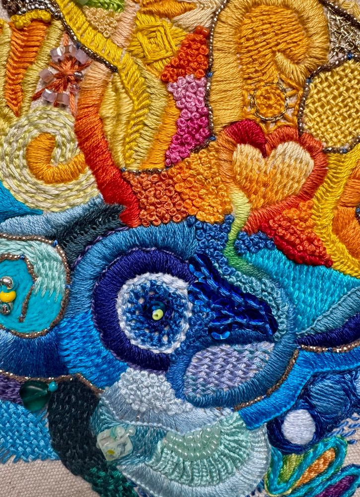 Close up of hand embroidery - yellows and oranges on top with blue tones on bottom. There is a huge variety of stitches and colors within the primary tones.