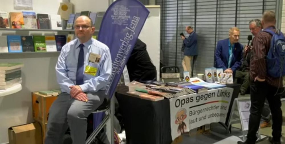 Axel Robert Göhring sitzt am Stand von EIKE. Daneben ist ein Stand von „Opas gegen Links“ zu sehen
